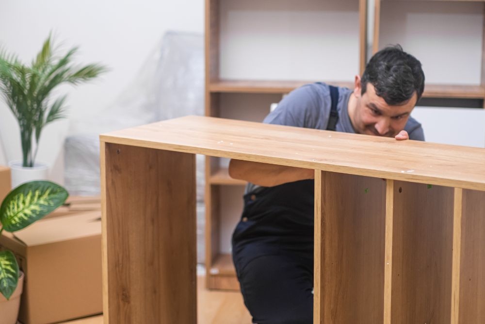 A man diligently works at a wooden table in a bright room, with tools and materials scattered around him.