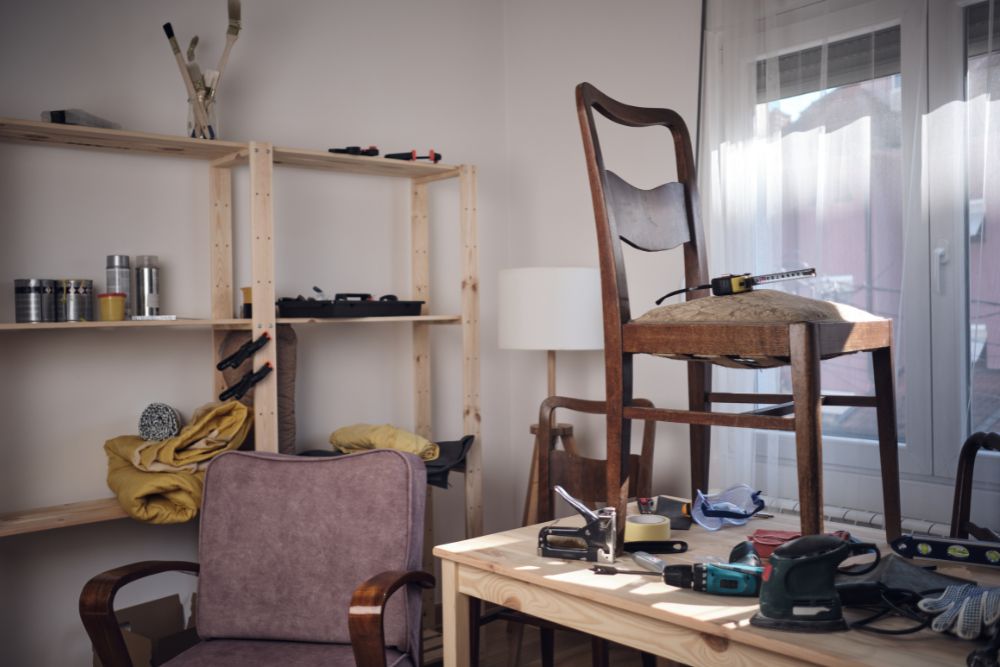 A cluttered workshop featuring a wooden chair on a table, tools, and supplies scattered across shelves and surfaces.