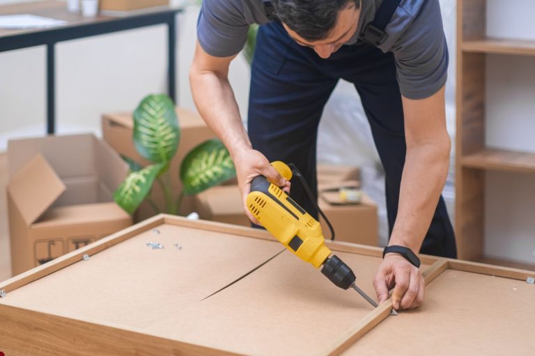 A man operates a drill while repairing a piece of furniture in a workshop setting.